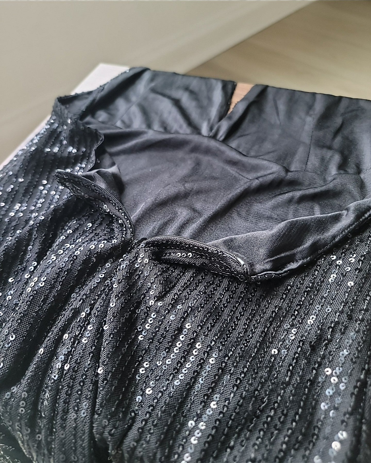 Close-up view of the back interior of a black sequin dress laid flat. A concealed black zipper runs vertically down the center of the back. The outer fabric is covered in small black sequins arranged in vertical rows, while the inside lining is smooth and matte. The image shows the seam construction and how the zipper is integrated into the back of the dress.