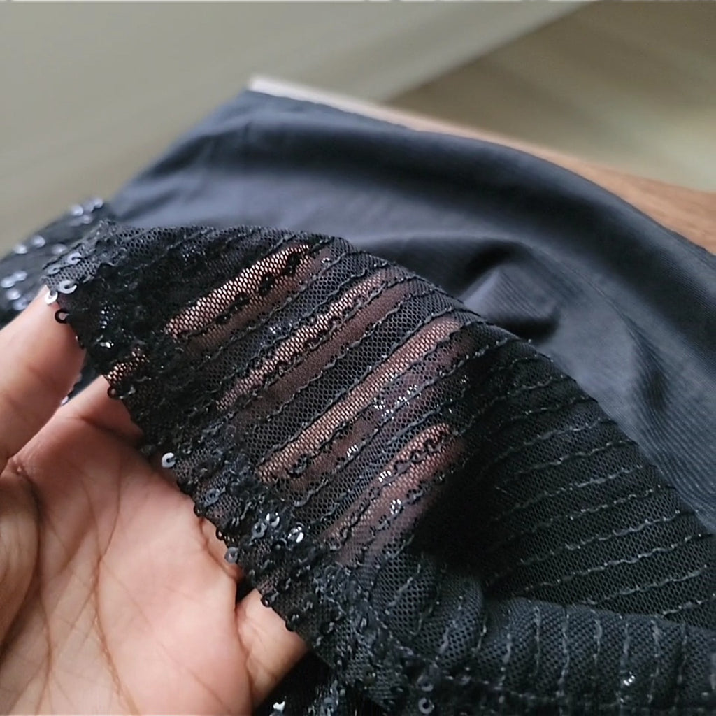 Close-up of the fabric of a black dress held in a hand, showing two distinct layers. The top layer is a sheer black mesh covered with small black sequins arranged in horizontal rows, allowing light to pass through. Beneath it is a smooth, opaque black satin lining that provides coverage. The image highlights the contrast between the textured, sheer sequin overlay and the soft satin underlayer.