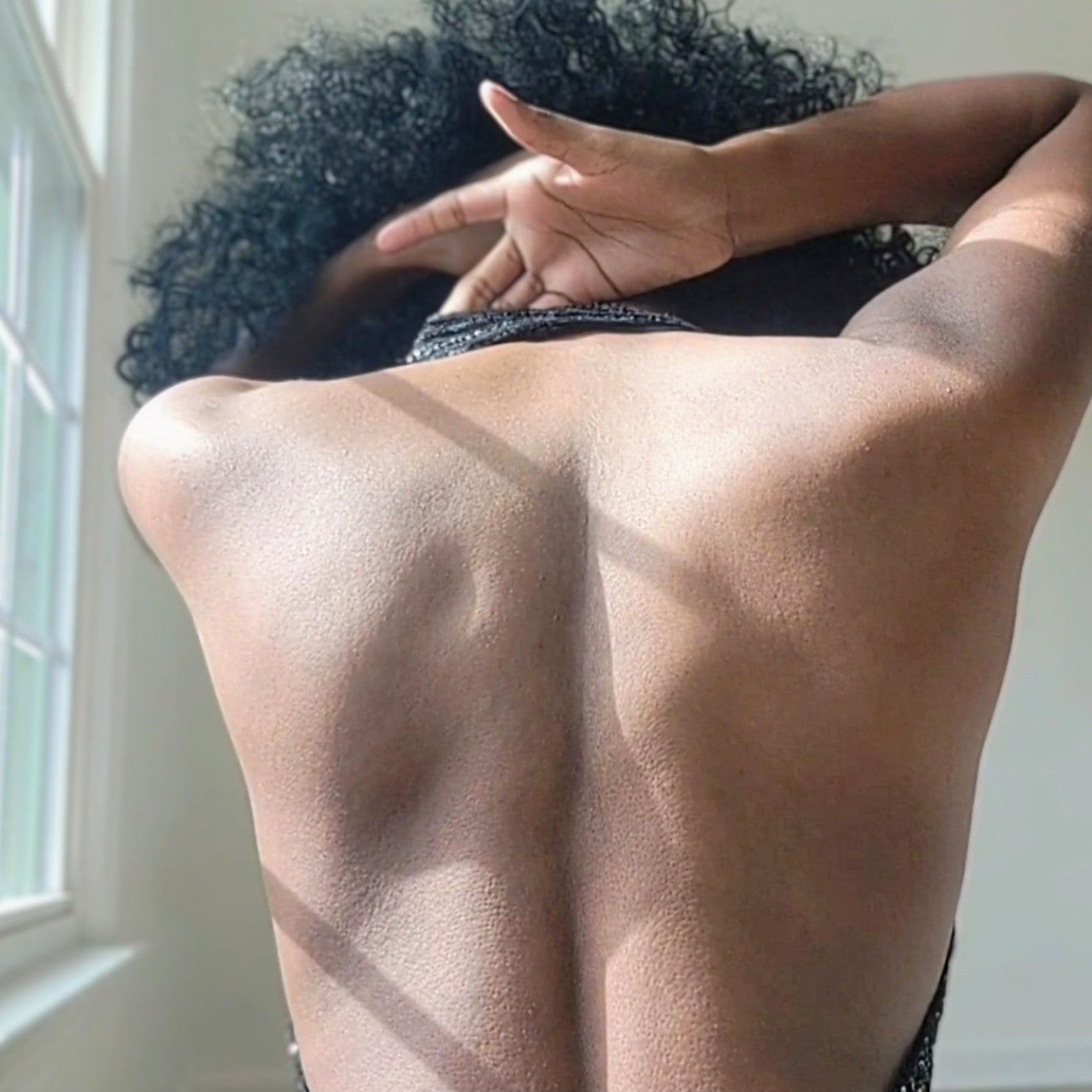 Close-up back view of a person wearing a black sequin maxi dress with a halter neckline. The halter strap wraps around the neck, leaving the upper back fully exposed. The dress fits closely along the back and torso, with vertical rows of small black sequins that reflect light subtly. The person’s arm is raised near the neck, and natural light from a nearby window highlights the texture of the fabric and the open-back design.