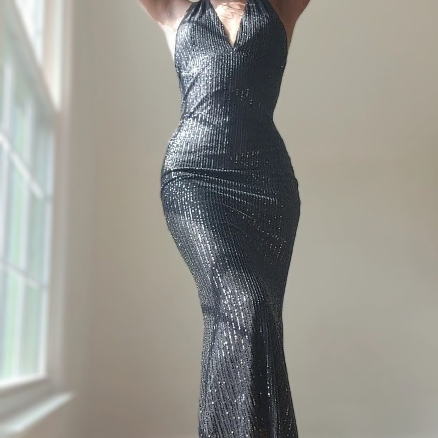 A person stands in soft natural light wearing a black, floor-length sequin maxi dress with a halter neckline. The dress features a deep V front and a fitted silhouette that contours closely through the waist and hips before falling straight to the hem. Vertical rows of sequins catch the light, creating a subtle shimmer across the fabric. The person’s arms are raised out of frame, emphasizing the neckline and upper bodice. A large window on the left allows daylight to illuminate the dress, while the backgrou