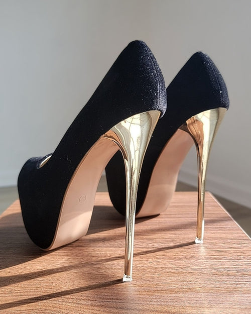 Back view of a pair of high-heeled platform shoes displayed on a wooden pedestal. The uppers are black velvet with a soft, matte texture. The heels are tall, thin, and metallic gold, reflecting light. The soles are a light neutral color. The shoes are positioned side by side in natural light, clearly showing the contrast between the black velvet material and the gold heels.