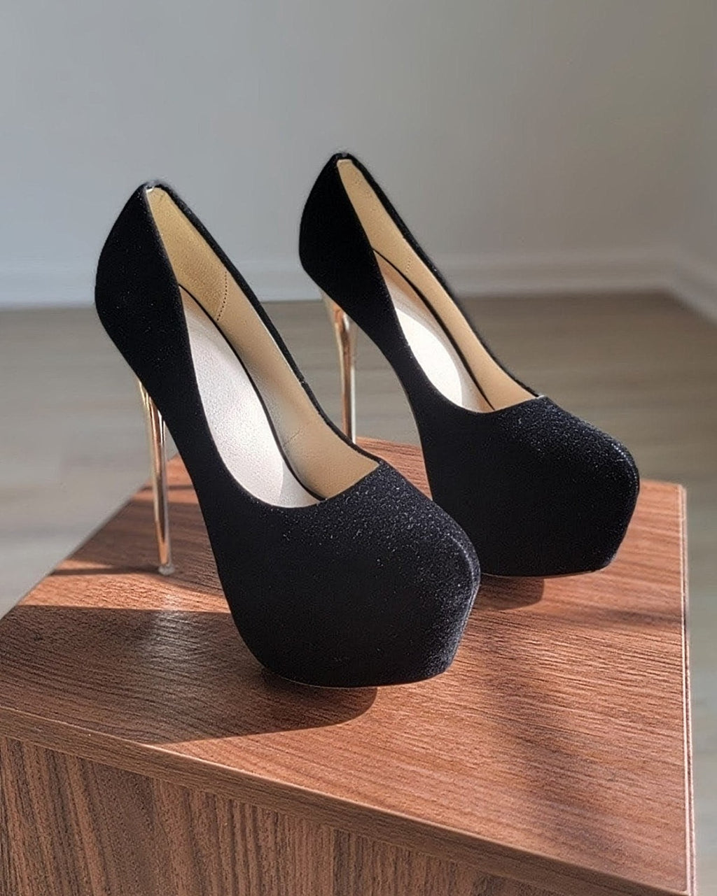 Pair of black velvet platform high-heeled shoes displayed on a wooden pedestal. The shoes have closed, rounded toes and tall, thin gold metallic heels. The interior lining is a light neutral color. The shoes are positioned side by side in natural light, clearly showing the platform sole, gold heel, and smooth velvet texture.